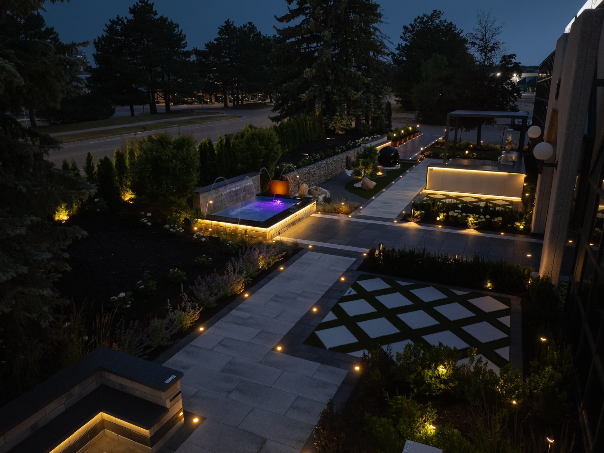 Aerial view at dusk of a landscaped property with lit walkways, terraces, square pavers in the lawn and a glowing spa.